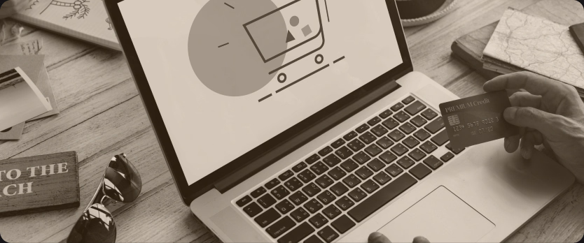 Laptop z ikoną koszyka zakupowego na ekranie, dłonie trzymające kartę płatniczą nad klawiaturą, obok filiżanka kawy i okulary przeciwsłoneczne